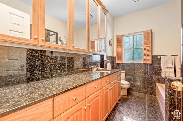 Bathroom with granite countertop, wooden cabinets, mirror, window with shutters, and bathtub with towel rack.