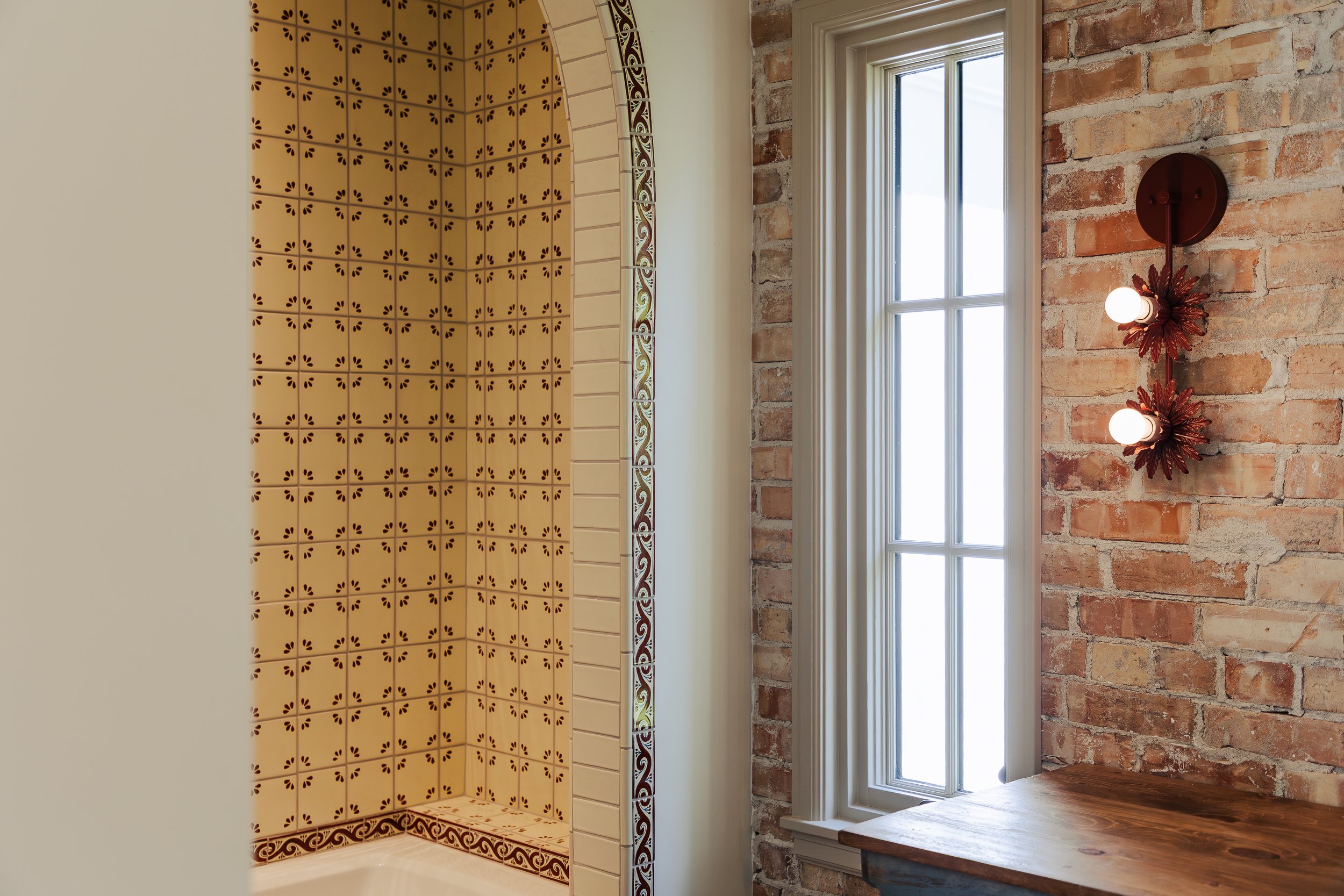 A bathroom with a shower area on the left, featuring yellow tiles with small brown floral patterns and decorative trim, next to a window with white framing and a brick wall with a red wall-mounted light fixture with two bulbs on the right.