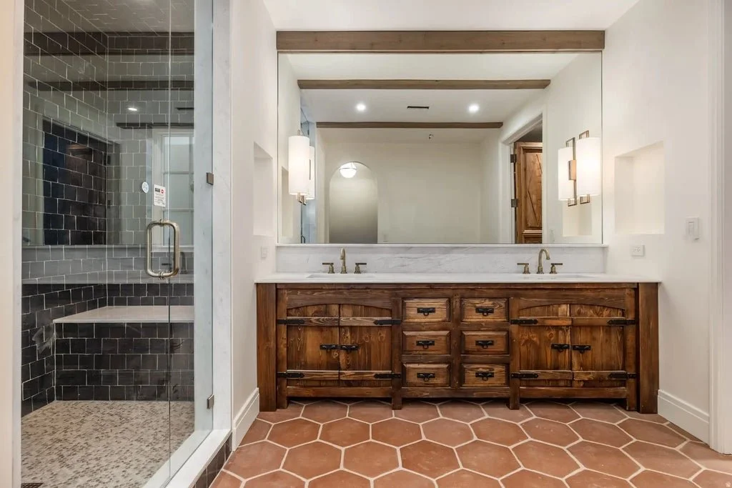 Bathroom with double sink vanity, large mirror, wall-mounted lights, and a tiled walk-in shower with black tiles, glass door, and pebble floor.