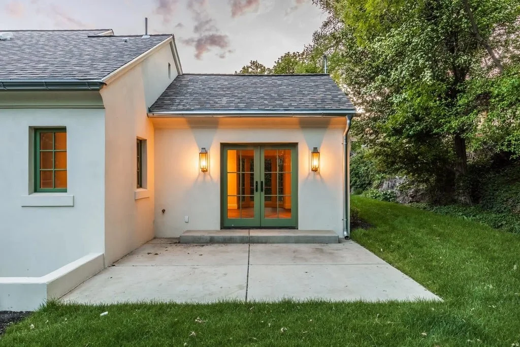 Backyard patio with concrete slab, white house exterior, green framed glass doors, two outdoor wall lanterns, grassy lawn, and trees in the background.
