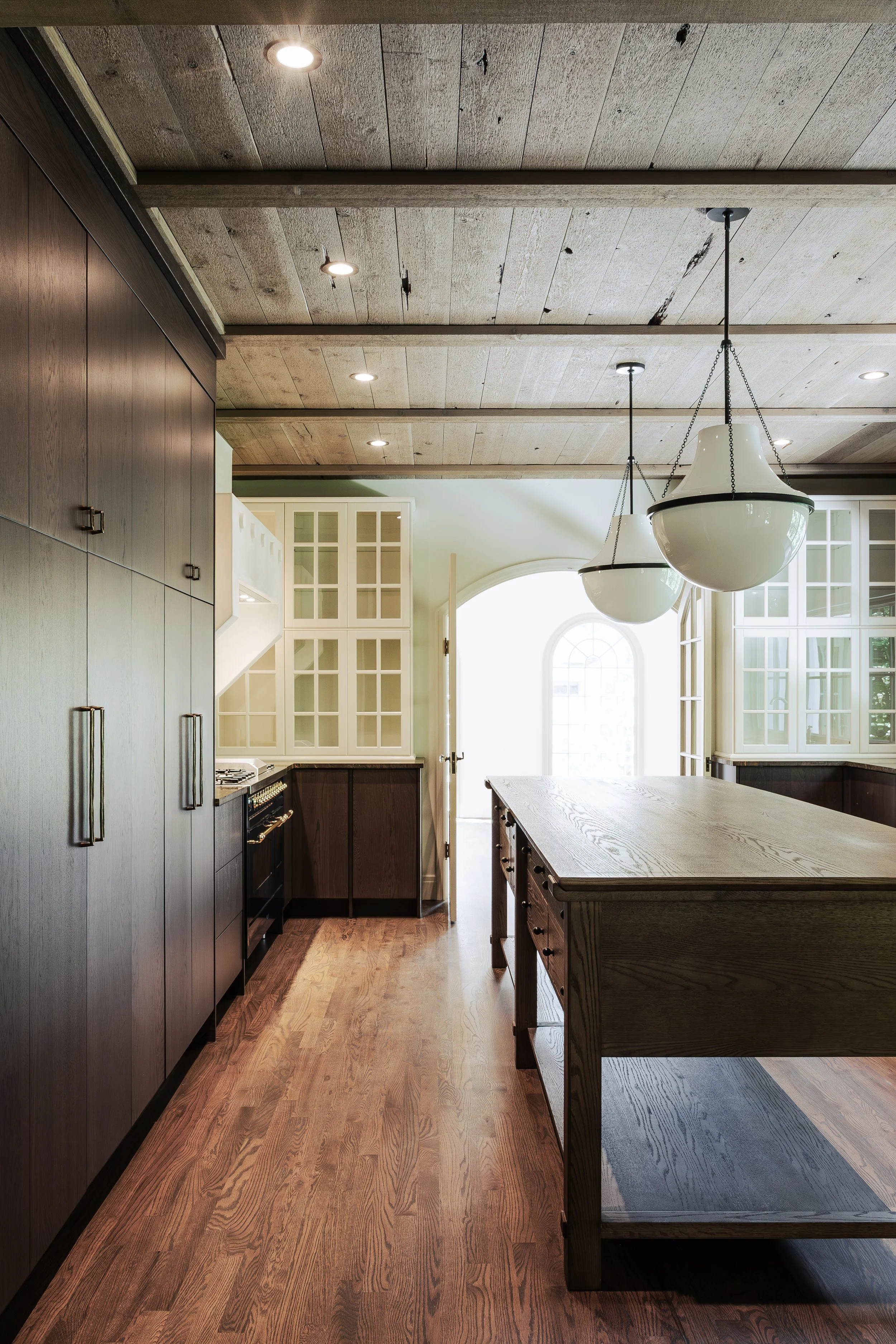 This image shows a modern kitchen with dark and light cabinets, a wooden kitchen island, a large window at the back, and a wood-paneled ceiling with recessed lighting and hanging pendant lights.