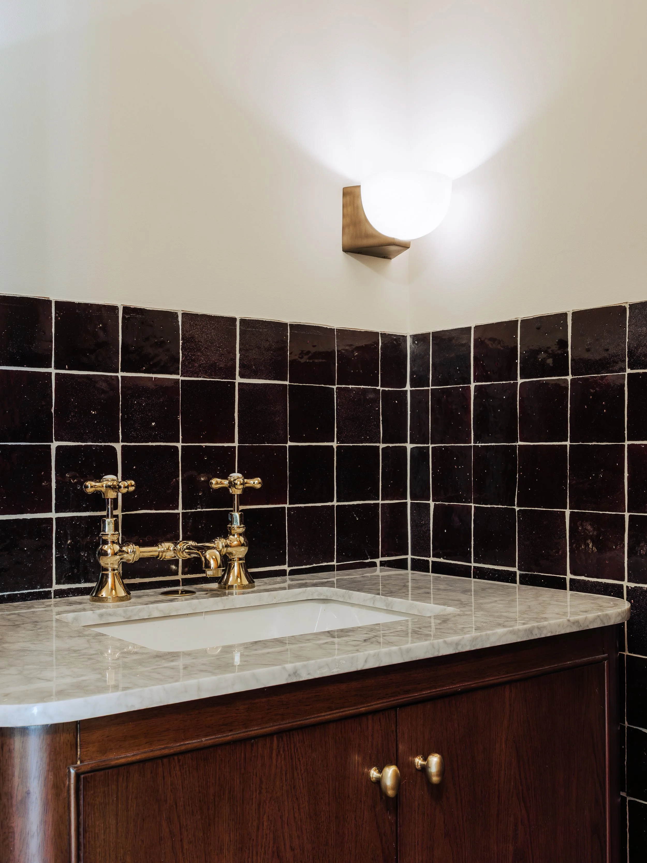 A bathroom vanity with a marble countertop, dark wood cabinet, and gold fixtures. A wall-mounted light fixture with a white spherical shade is above the mirror, illuminating the wall.