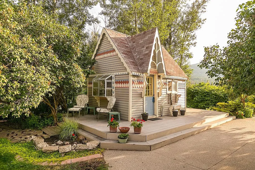 Small cottage with decorative trim, a small porch, and outdoor seating, surrounded by plants and trees.