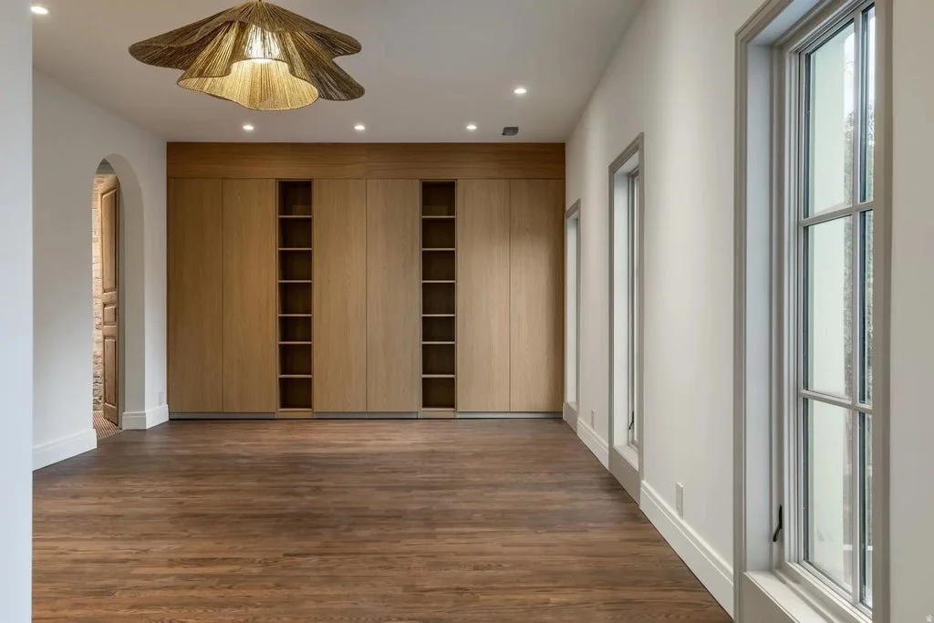 Empty room with hardwood floors, white walls, and large windows. A closet with wooden sliding doors and open shelves is visible. A decorative ceiling light fixture is also seen.