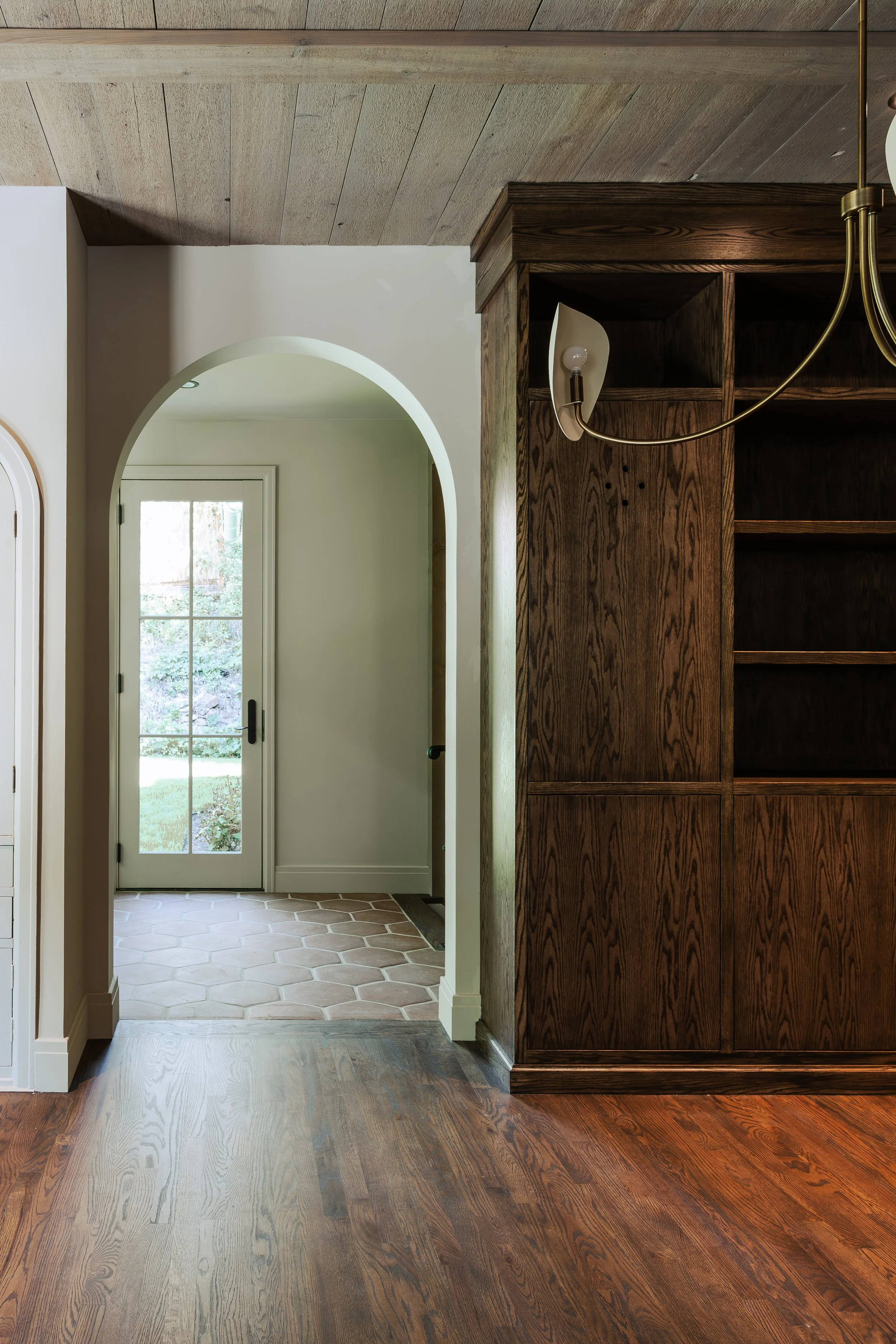 Interior view of a home with wooden ceiling, dark wood shelving unit, and two-tone flooring; stone tile in the entryway, hardwood in the main area, with a white door leading outside.
