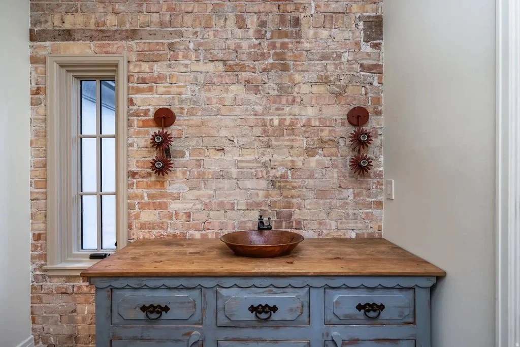 Rustic bathroom vanity with a wooden countertop, a wooden sink, and a distressed blue base cabinet, set against an exposed brick wall with decorative wall sconces and a small window.