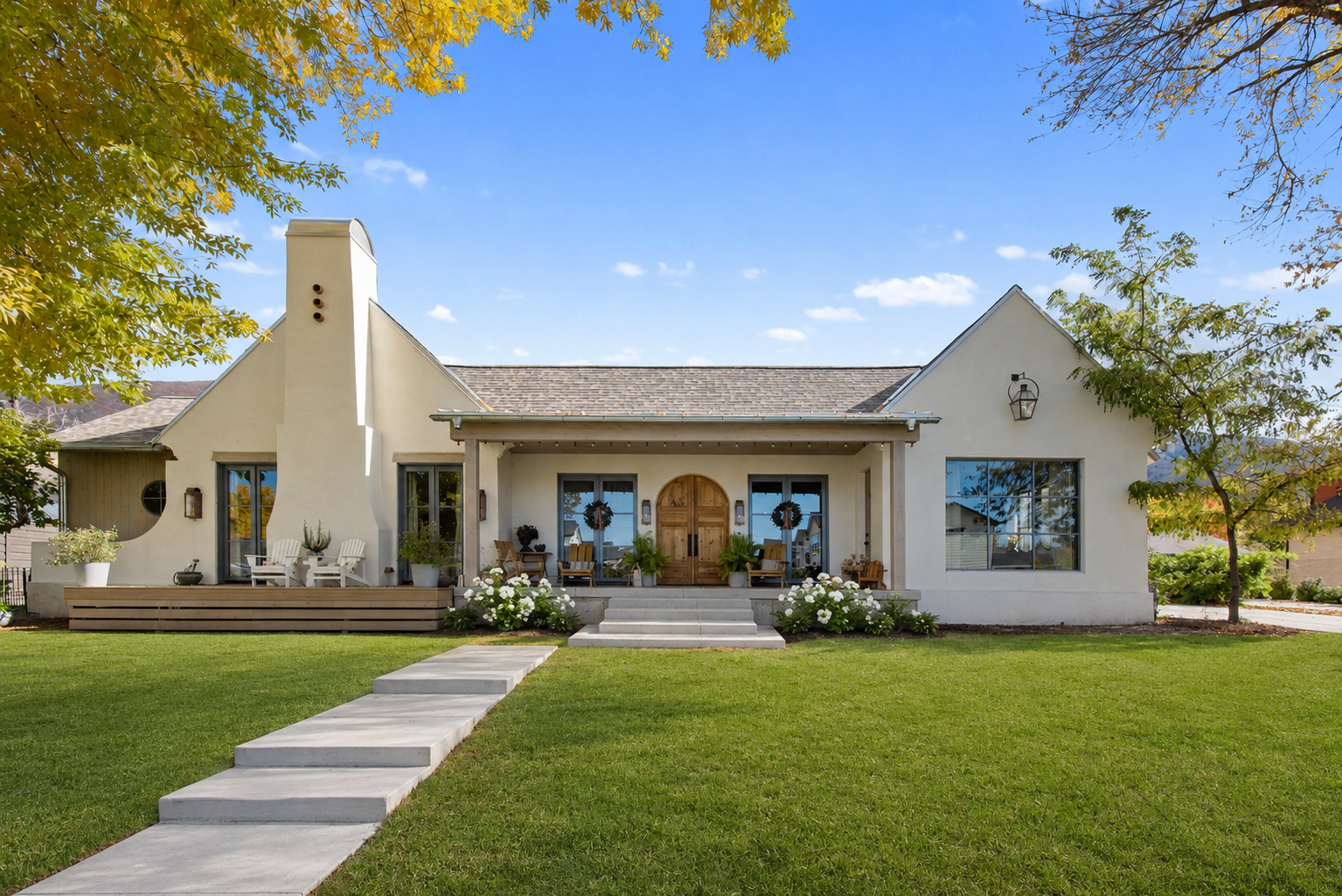 A modern white house with a pitched roof, large windows, and a front porch decorated with wreaths and plants, surrounded by a green lawn and trees under a blue sky.