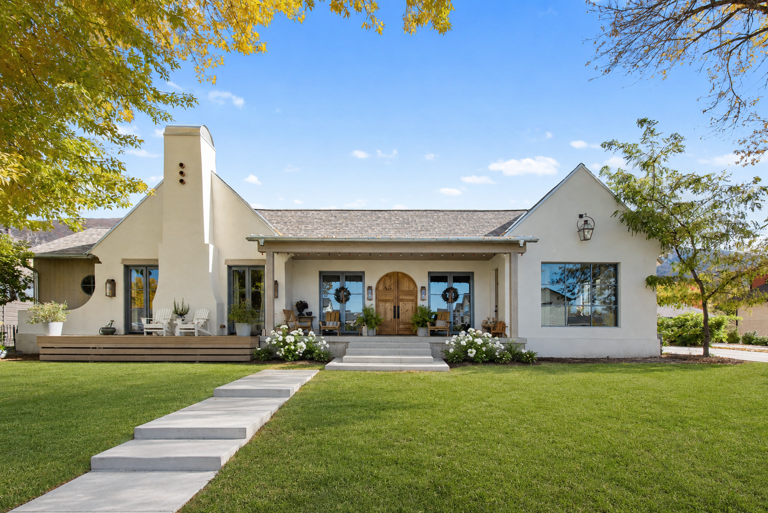 A modern house with a white stucco exterior, a covered front porch, and a well-maintained lawn with stepping stones leading to the entrance. There are trees with yellow and green leaves on either side of the house, and the sky is clear with a few clouds.
