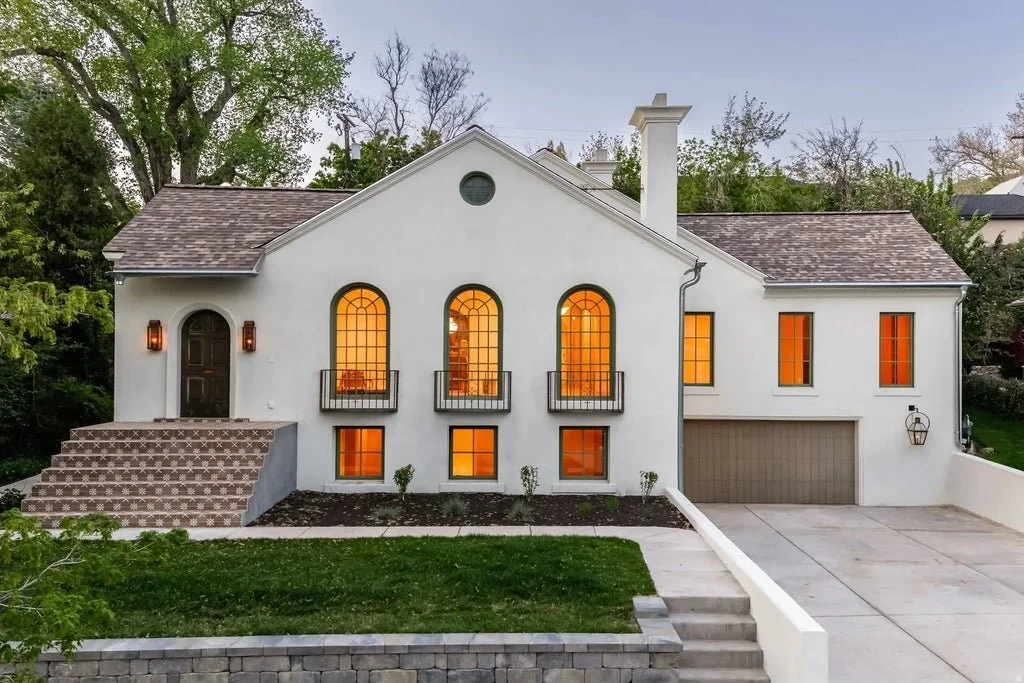 A white two-story house with arched windows, a brown front door, and a garage. The house has a tiled front steps and a small lawn with a few bushes. Lights are on inside the house, glowing through the windows.