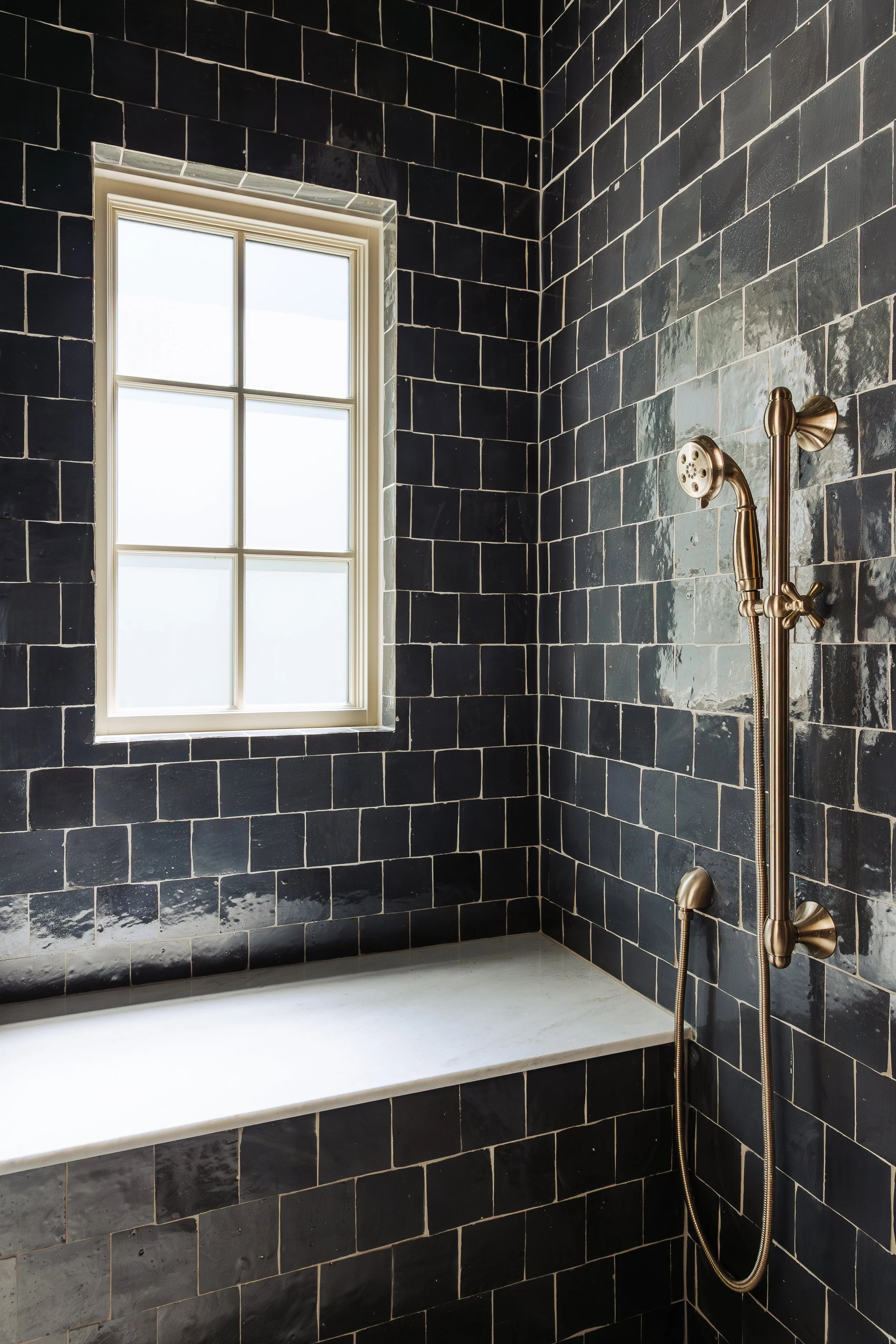 A bathroom shower area with black tiled walls, a window with white trim, a built-in white bathtub, and a gold shower fixture. The showerhead is mounted on the wall with a hose hanging down.