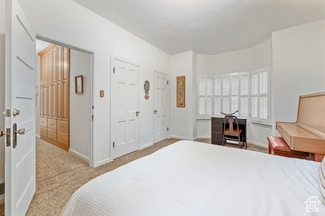 A bedroom with white walls, a bed with white bedding, a wooden piano with a stool, a desk near a large bay window with closed blinds, and a hallway with closet doors and a wooden cabinet.