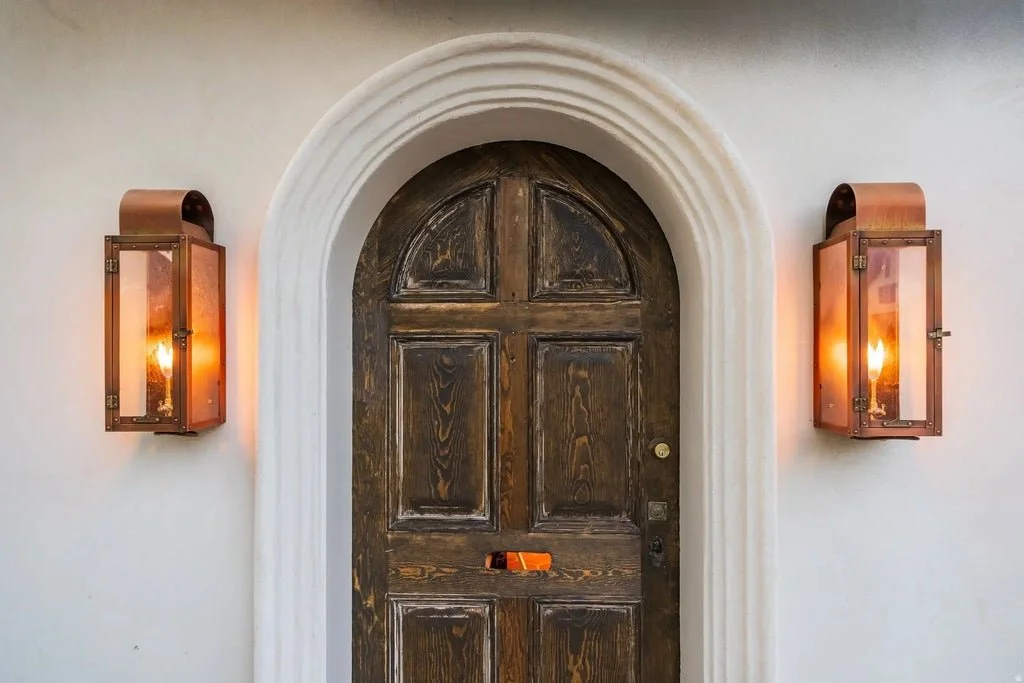 Front door with a rounded white archway, two wall-mounted copper and glass lantern-style lights on each side, and a dark wood door with a mail slot.