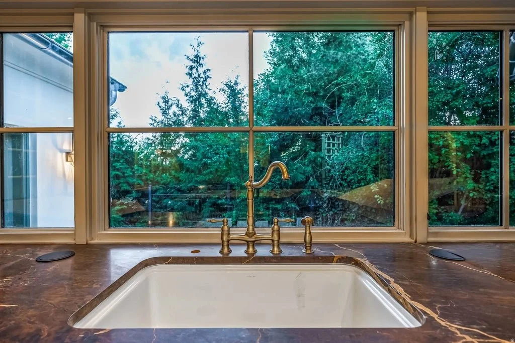 Kitchen sink with brass faucet and handles on a marble countertop, large windows showing green trees outside.