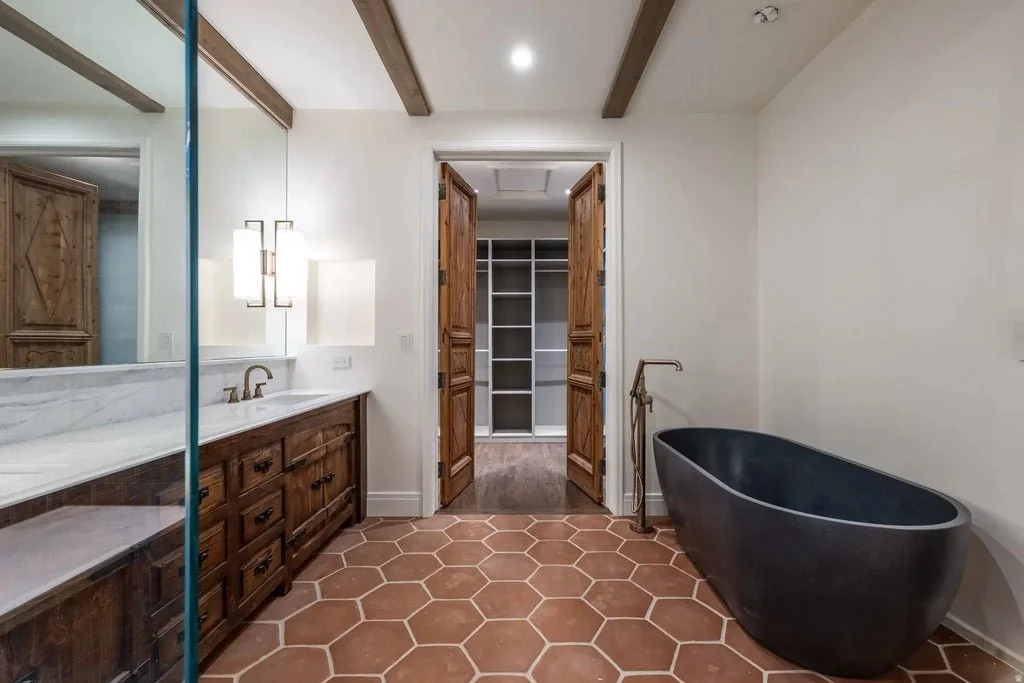 Bathroom with hexagonal terracotta tile flooring, a black freestanding bathtub, wooden double doors leading to a walk-in closet, and a wooden vanity with a marble countertop and two sinks.
