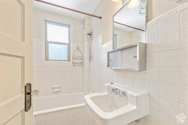 Small bathroom with a bathtub, a window, a wall-mounted ceramic sink, a mirrored medicine cabinet, and a showerhead, all in a neutral color scheme.