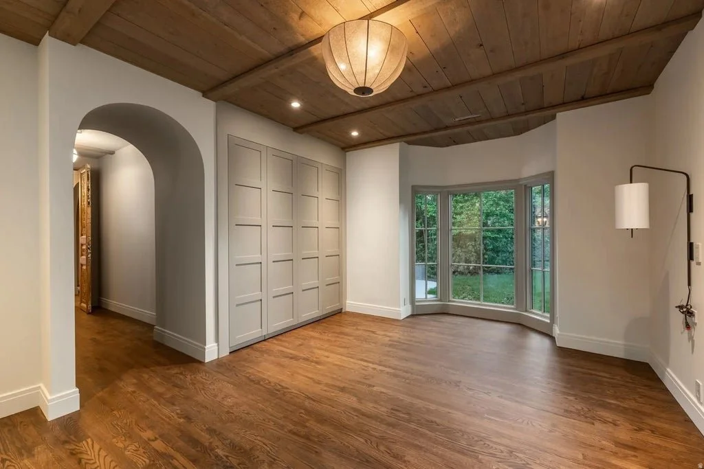 Empty living room with wooden ceiling and floor, gray built-in closet, large bay window, and wall sconce lighting.