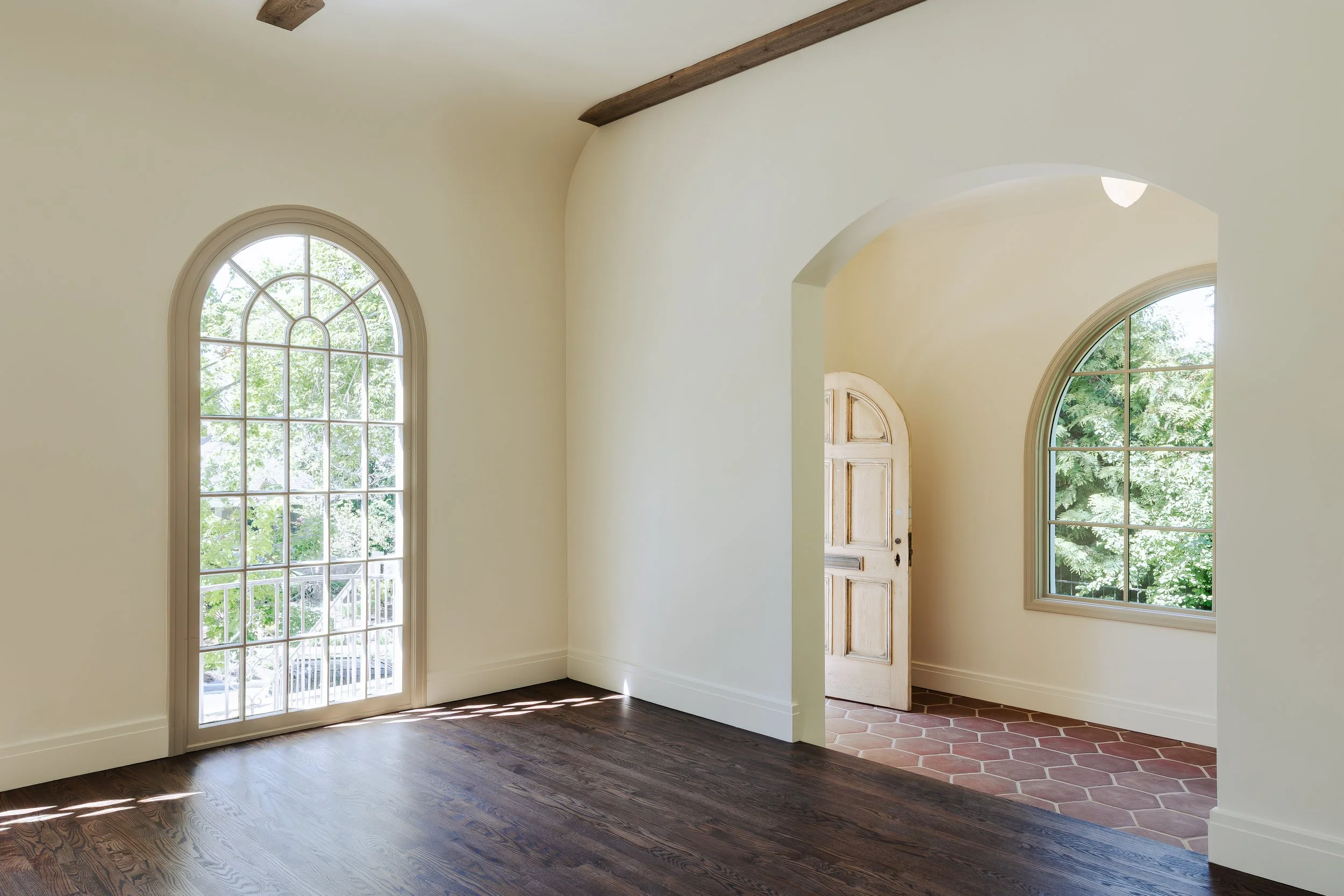 Interior view of a house with large arched windows, hardwood flooring, and a doorway with brick flooring leading outside.