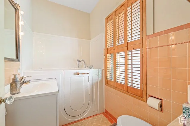 Bathroom with a washing machine, a window with wooden shutters, a toilet paper roll, and beige tiles.