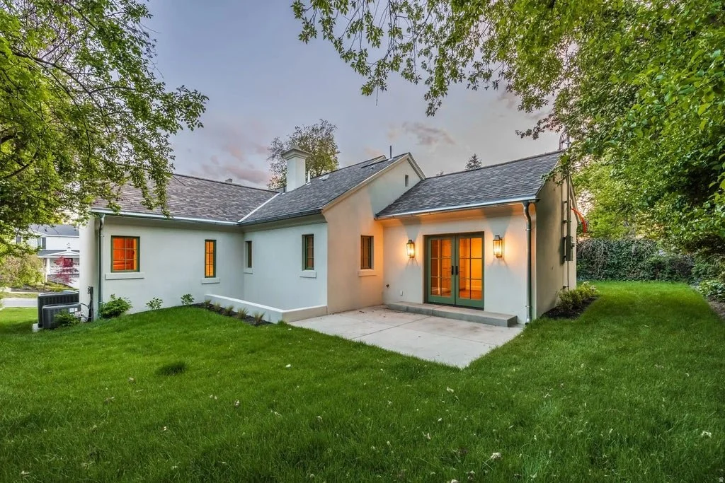 A house with white exterior walls, a gray shingle roof, and a small patio area with double glass doors illuminated by exterior lights. The house is surrounded by a well-maintained green lawn and trees.