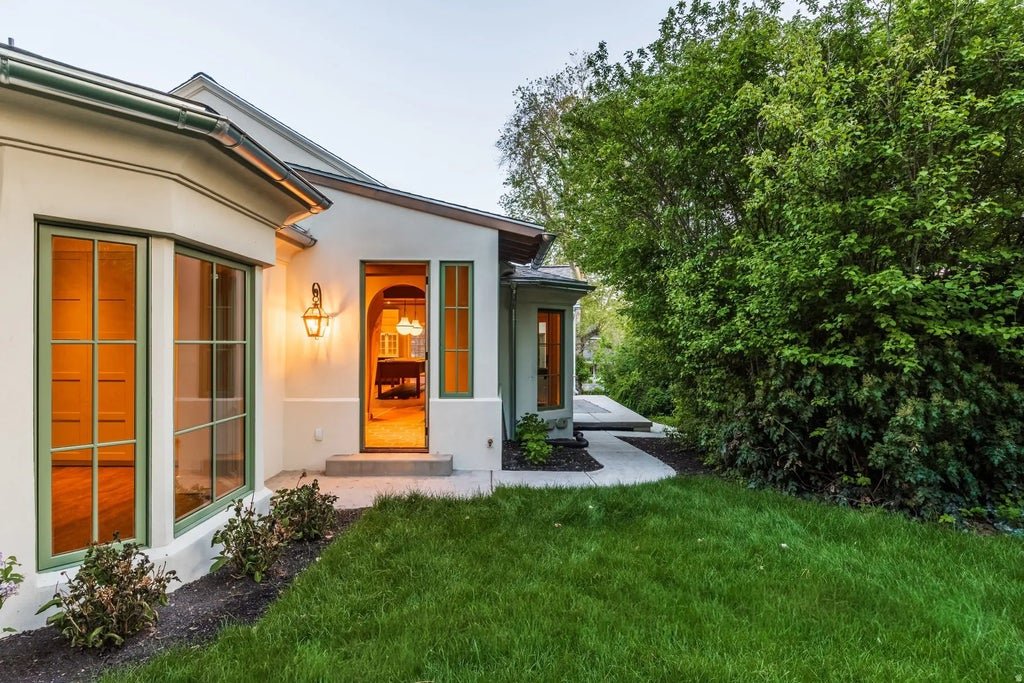 A house with large windows, a porch with a door, and outdoor lighting, surrounded by a green lawn and tall bushes.