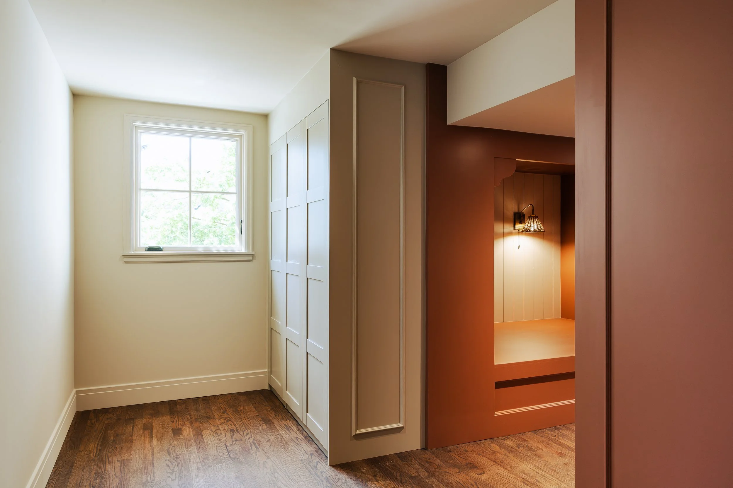 Bunk room with a window, white walls, and a shadowed hallway with wooden flooring.