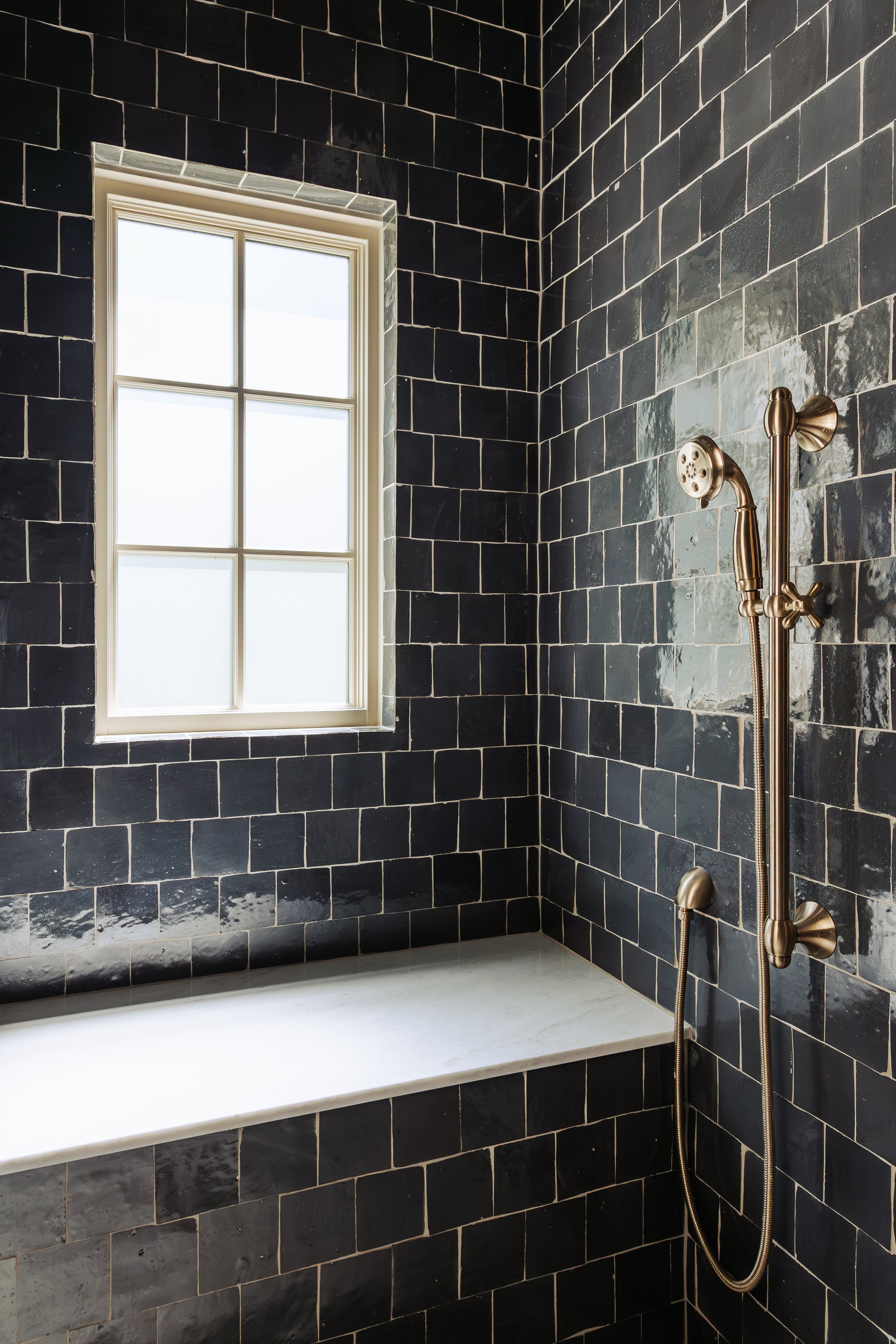 A bathroom shower area with black tiled walls, a white bathtub, and a vintage-style brass shower fixture. A window with white trim is visible on the tiled wall.