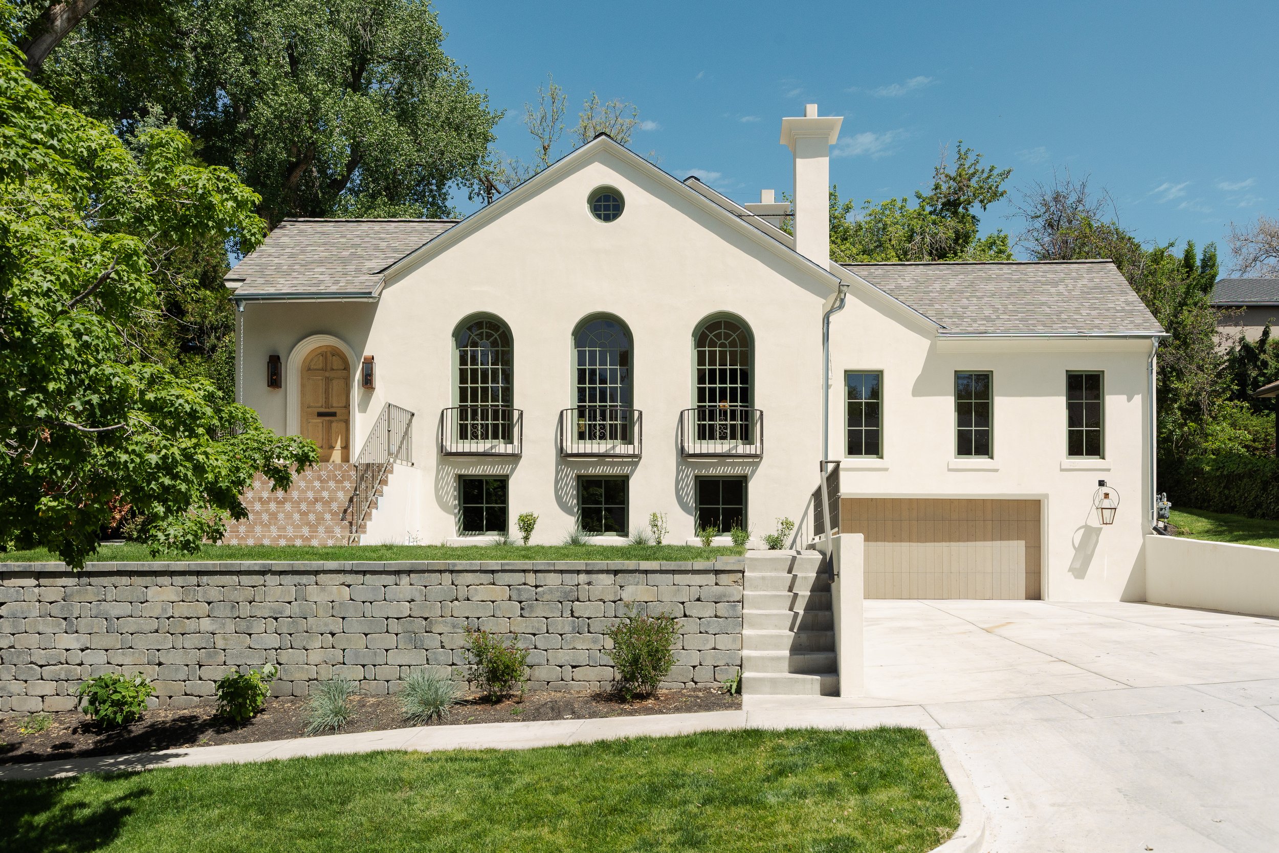 Front view of a white two-story house with arched and rectangular windows, a gray shingle roof, a chimney, front stairs, and a driveway, surrounded by greenery and trees under a clear blue sky.