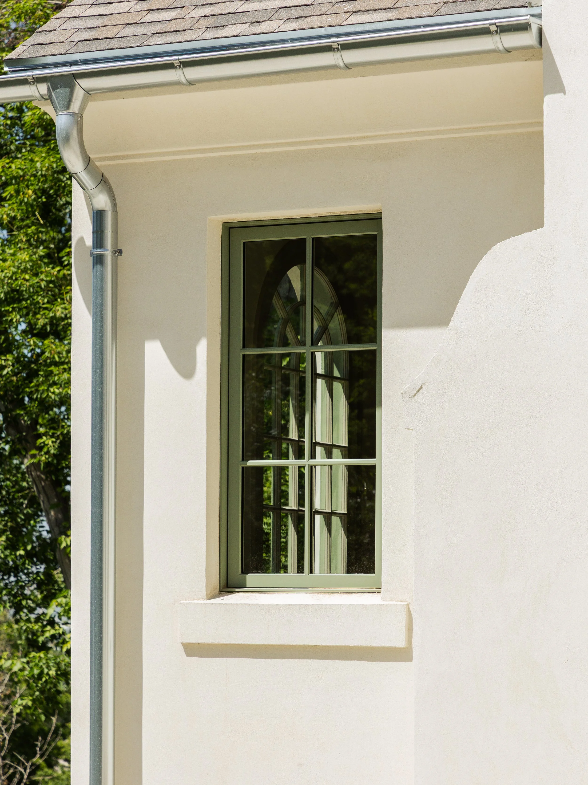 A section of a house exterior with a green-framed window with multiple small panes and a decorative arched top. The house has white stucco walls, a sloped gray roof, and a green tree with sunlight in the background.