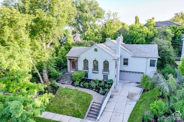 A white two-story house with a curved front staircase, surrounded by lush green trees and shrubs, with a concrete driveway leading to a garage.