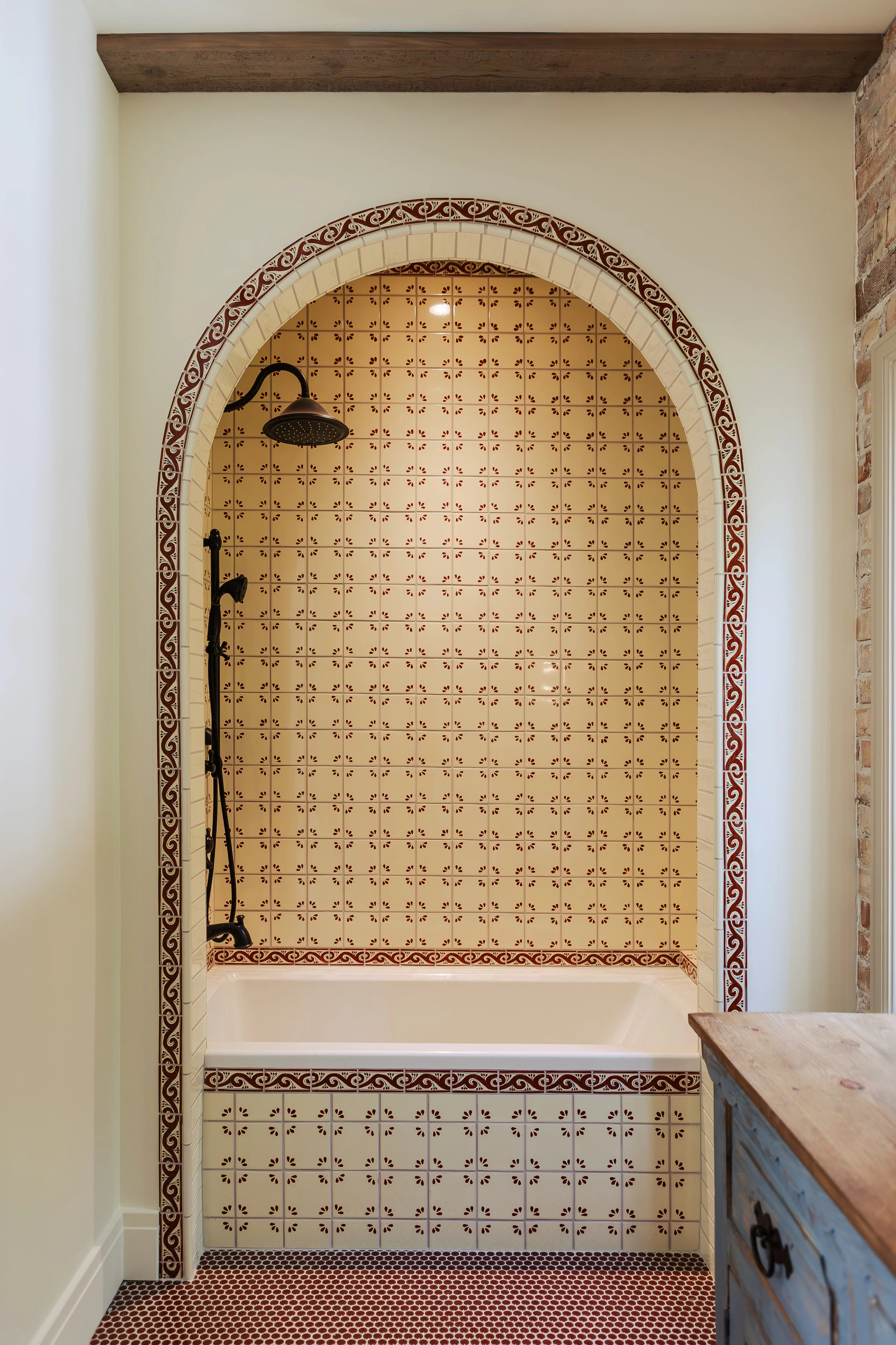 A walk-in shower with beige and red patterned tiles inside a bathroom. The shower has a black rainfall shower head and a brown wood beam ceiling. There is a brick wall on the right and a wooden cabinet in the lower right corner.