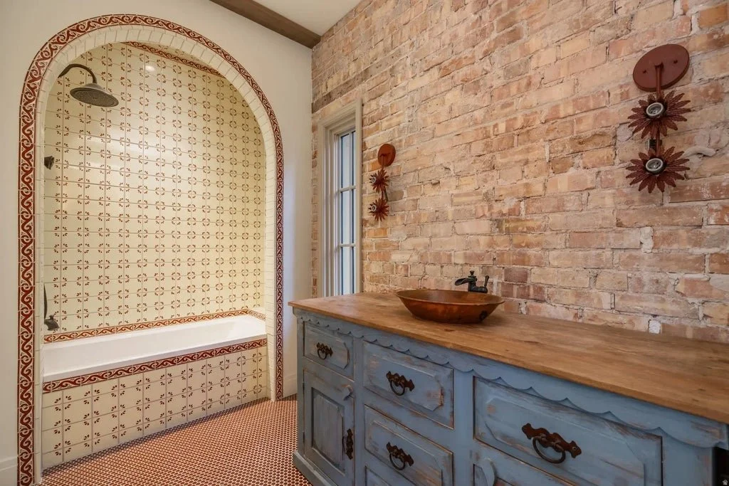 A bathroom with a brick wall, a blue wooden vanity with a wooden bowl sink and vintage hardware, and a shower area with patterned tiles and a rain shower head.