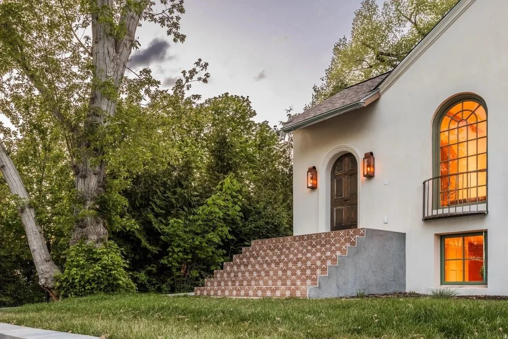 Exterior of a house with steps leading up to a wooden door, two wall-mounted lanterns, illuminated windows with a warm glow, and lush green trees in the background.