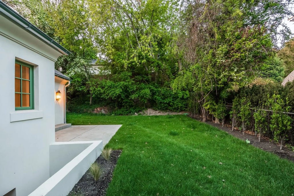 A backyard with a manicured green lawn, a white house with a small porch, a window with green trim, and a outdoor light fixture. There are trees and bushes along the fence line.