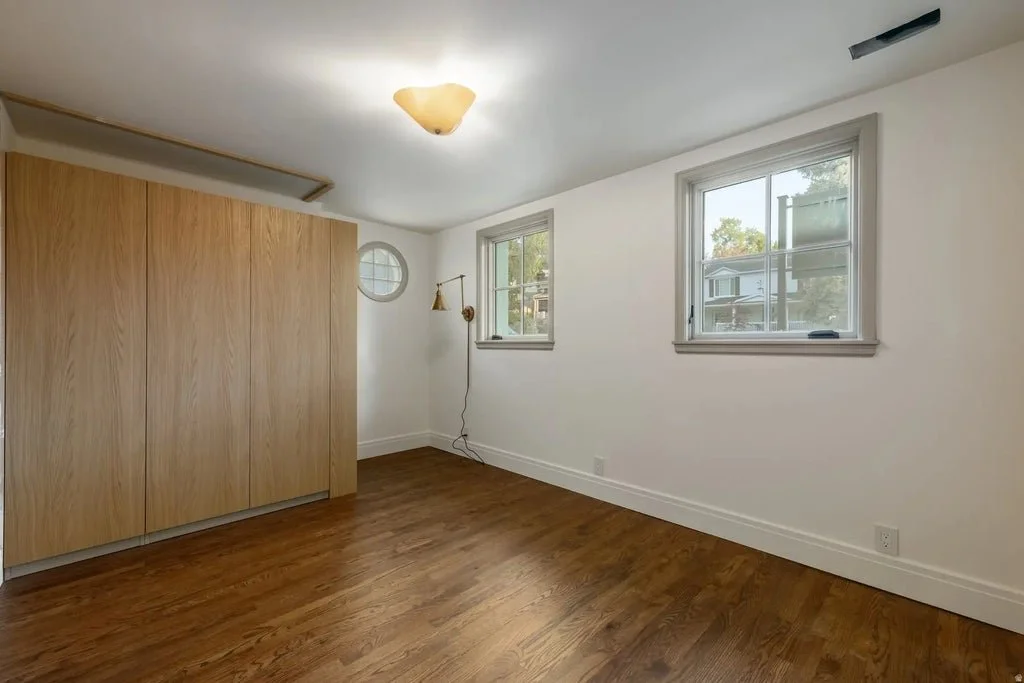 Empty room with hardwood flooring, white walls, two windows, a ceiling light fixture, and a wooden closet.