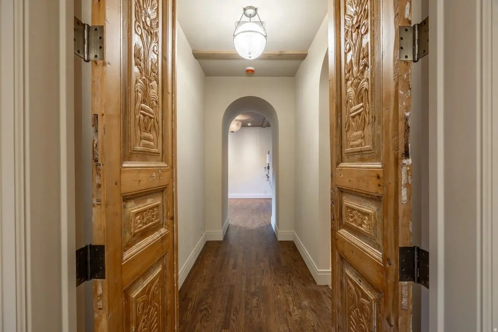 Wooden carved double doors opening into a hallway with white walls, hardwood floors, and an arched doorway leading into another room.