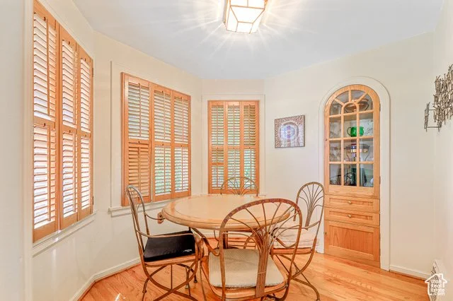 Dining area with a round wood table and four wooden chairs, three with cushions, in front of three windows with wooden shutters, and a built-in wooden cabinet on the right side, in a room with hardwood flooring and a ceiling light fixture.