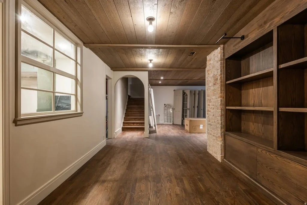 Interior of a home with hardwood floors, wooden ceiling, large window, staircase, brick wall, and built-in wooden shelves.