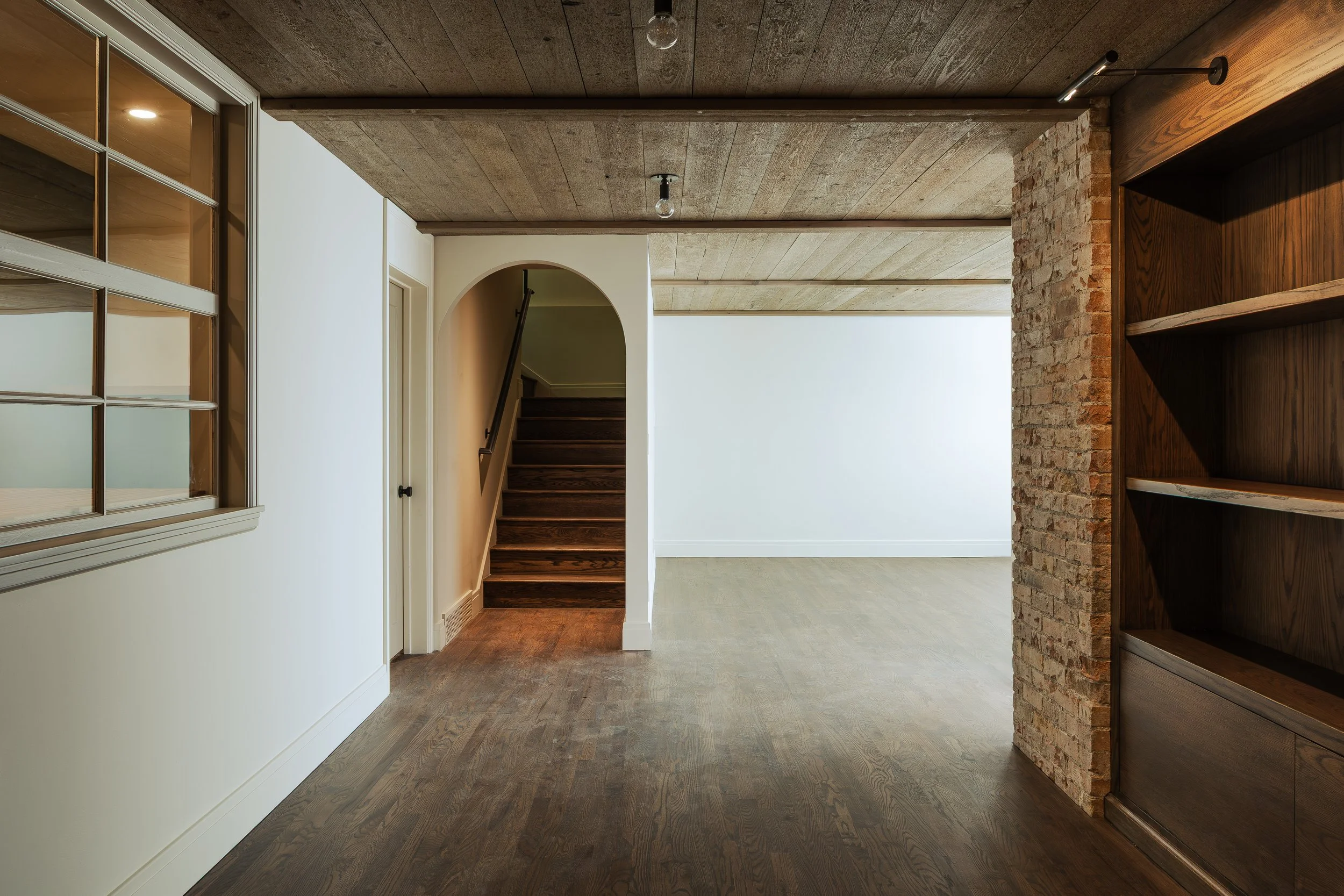 Interior of a modern home with a wooden staircase, brick wall, and wooden shelves, illuminated by ceiling lights.