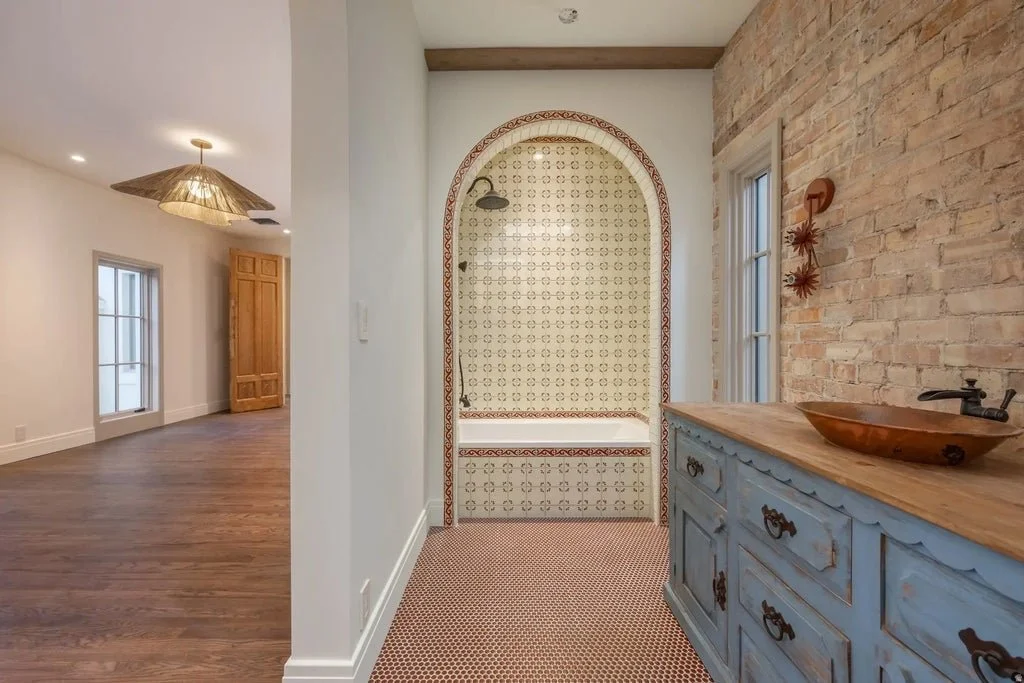 Interior view of a home with a hallway leading to a nook with a tiled wall, a potted plant, a window, and a rustic blue cabinet with a wooden bowl on top.