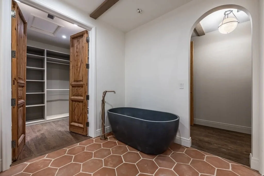 Bathroom with a black freestanding bathtub, wooden double doors leading to a walk-in closet, and an arched doorway with a ceiling light, terracotta hexagonal floor tiles, and white walls.