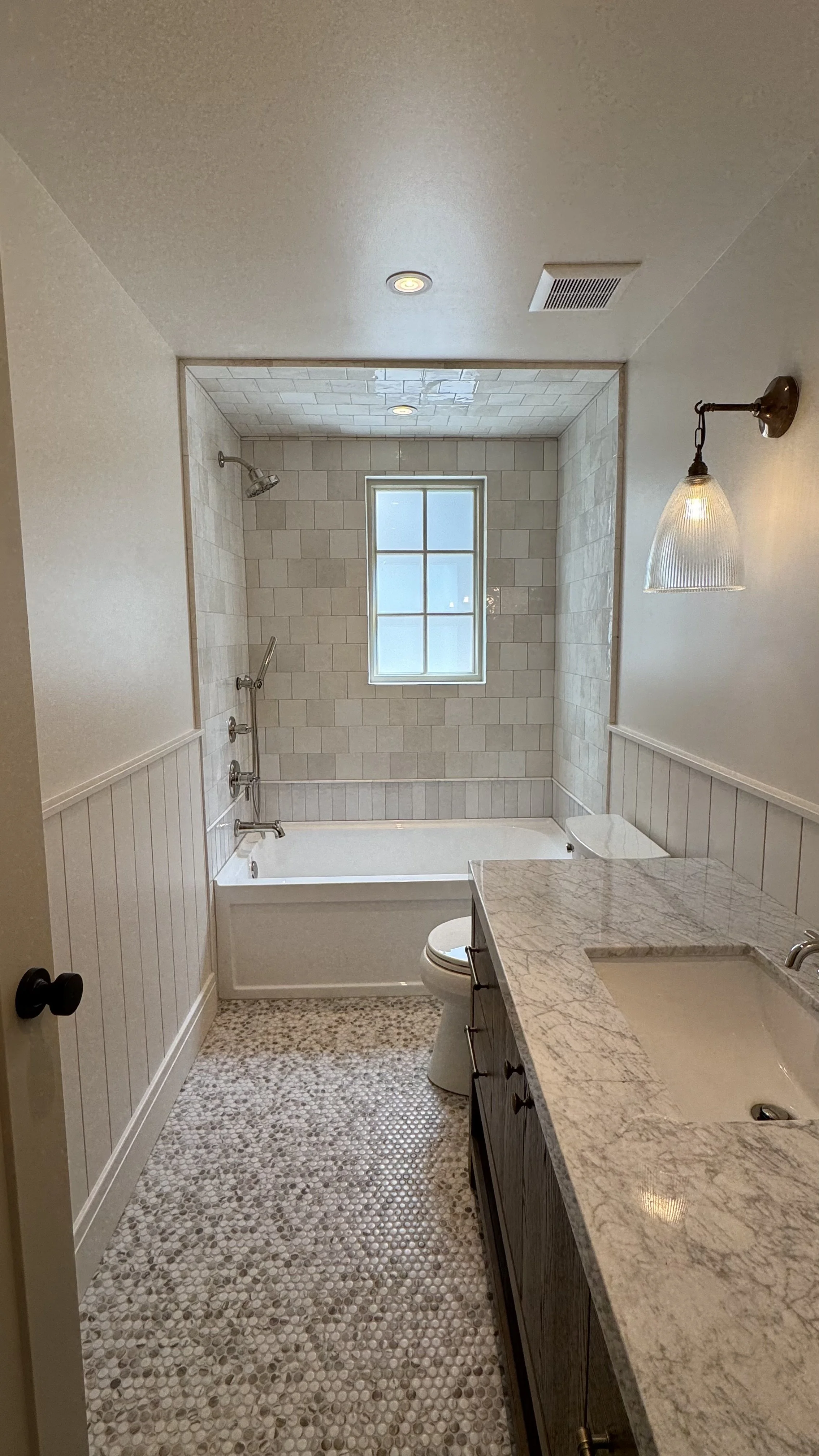 Bathroom with bathtub, tiled walls, window, toilet, marble countertop with sink, and textured pebble floor.