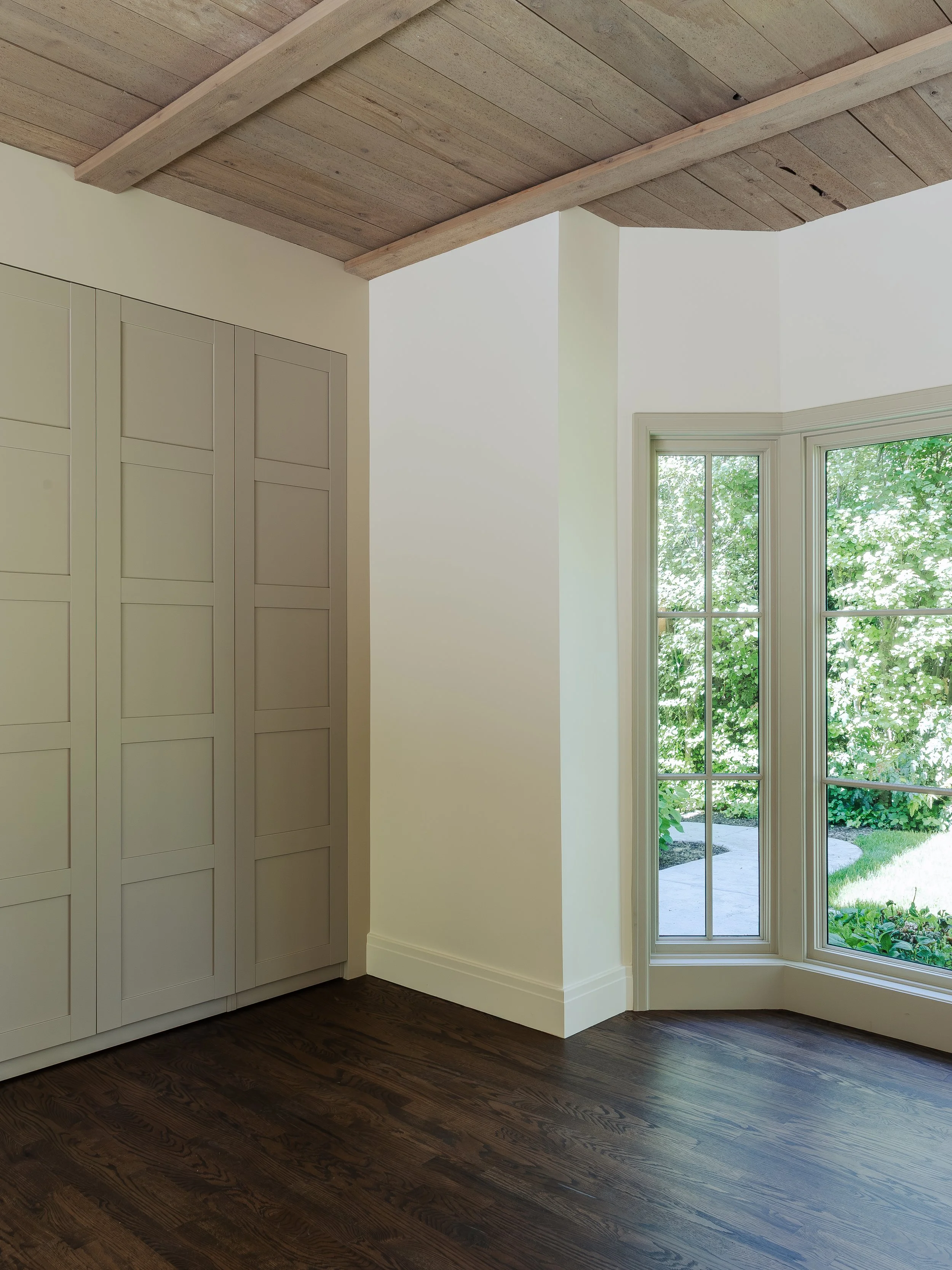 Interior view of a room with dark wooden flooring, white walls, a window with multiple panes through which green foliage is visible, and a built-in white cabinet with paneled doors on the left side.