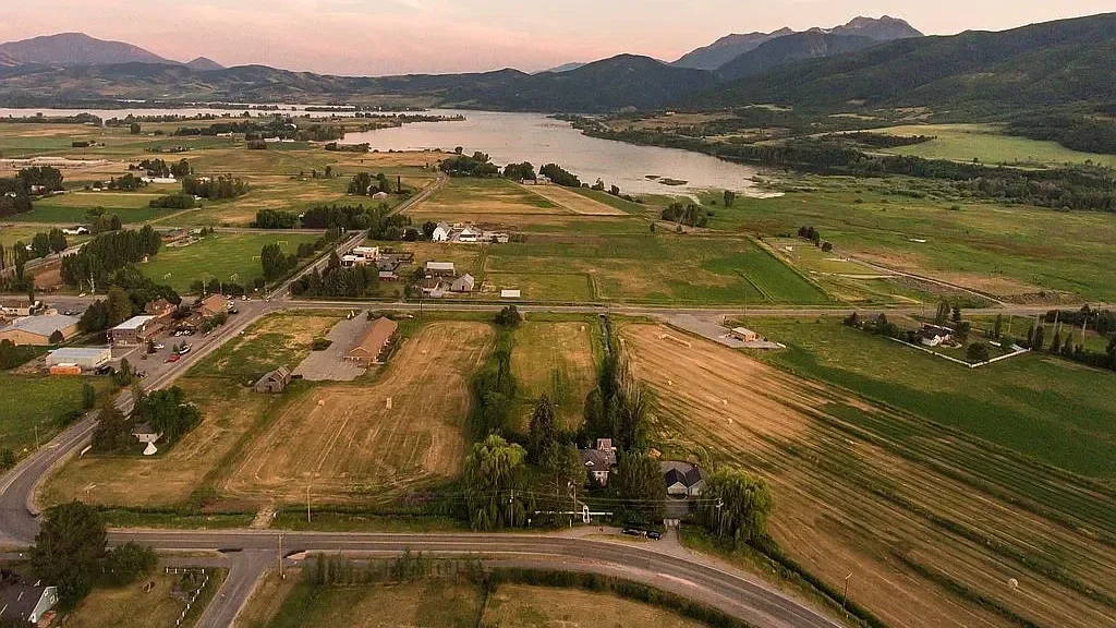 Aerial view of a rural landscape with fields, roads, houses, and a large river, with mountains in the background during sunset.