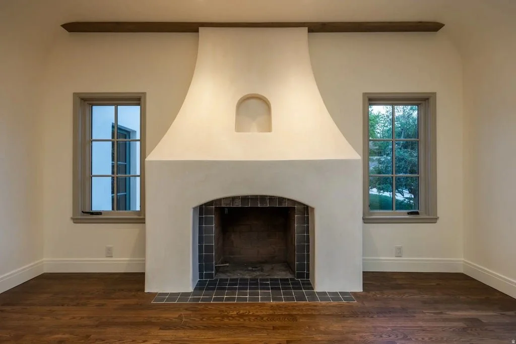 Living room with a white stucco fireplace, two windows on either side, and wooden flooring.