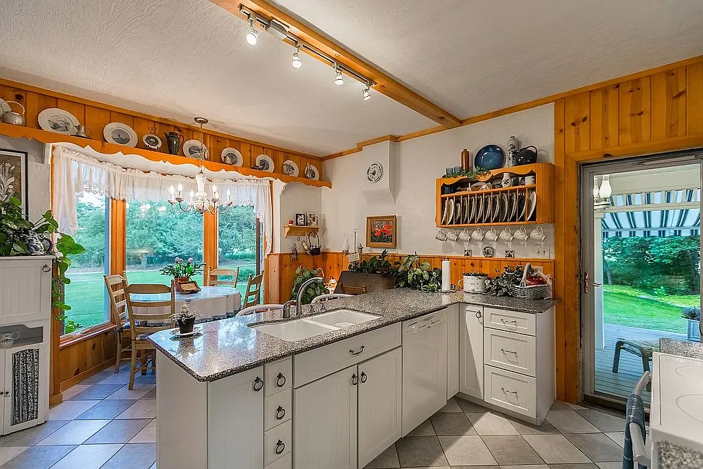 Kitchen with wooden and white cabinetry, granite countertops, and plenty of windows offering a view of a green lawn outside. There is a dining area with a table and chairs near the windows and decorative dishes and plates on shelves above.