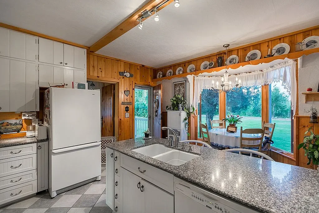 Kitchen with white cabinets, a granite countertop island, and a windowed dining area with a round table and chairs overlooking a green backyard.