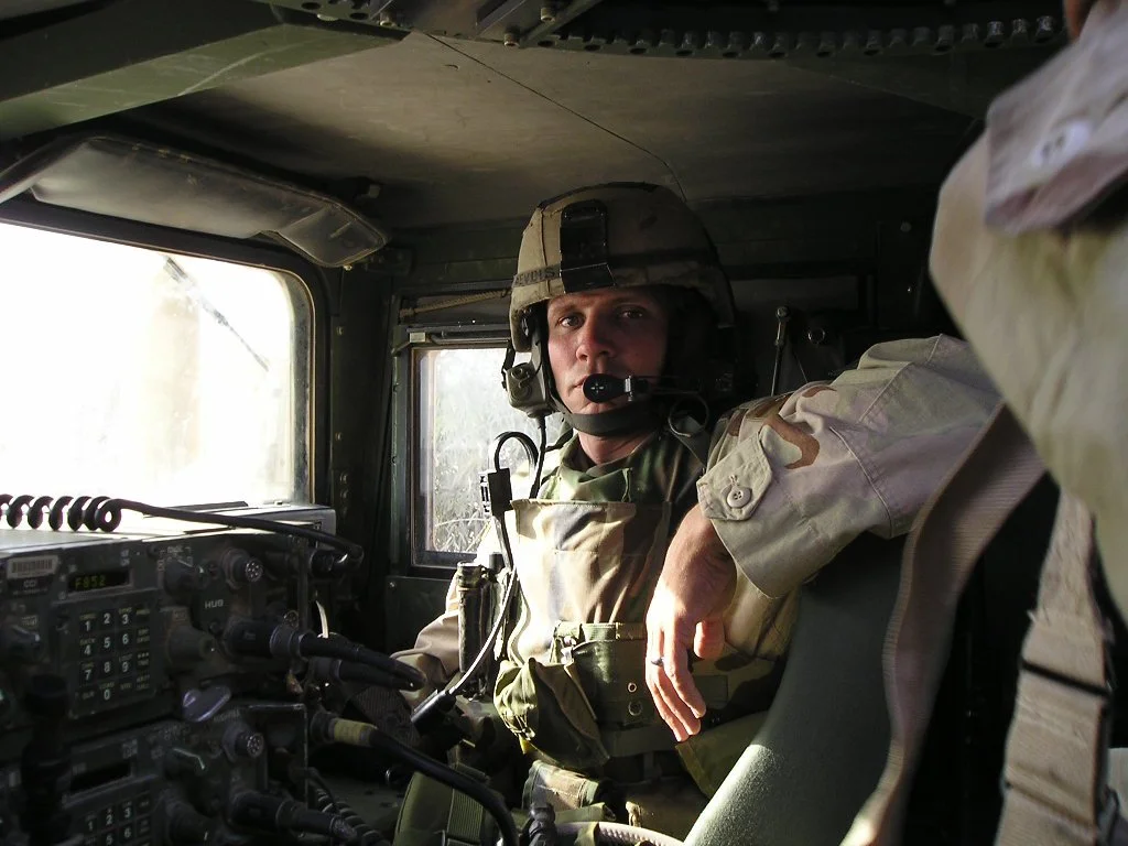 A soldier in military gear, including a helmet and camouflage uniform, sitting inside a military vehicle, looking out from the window.