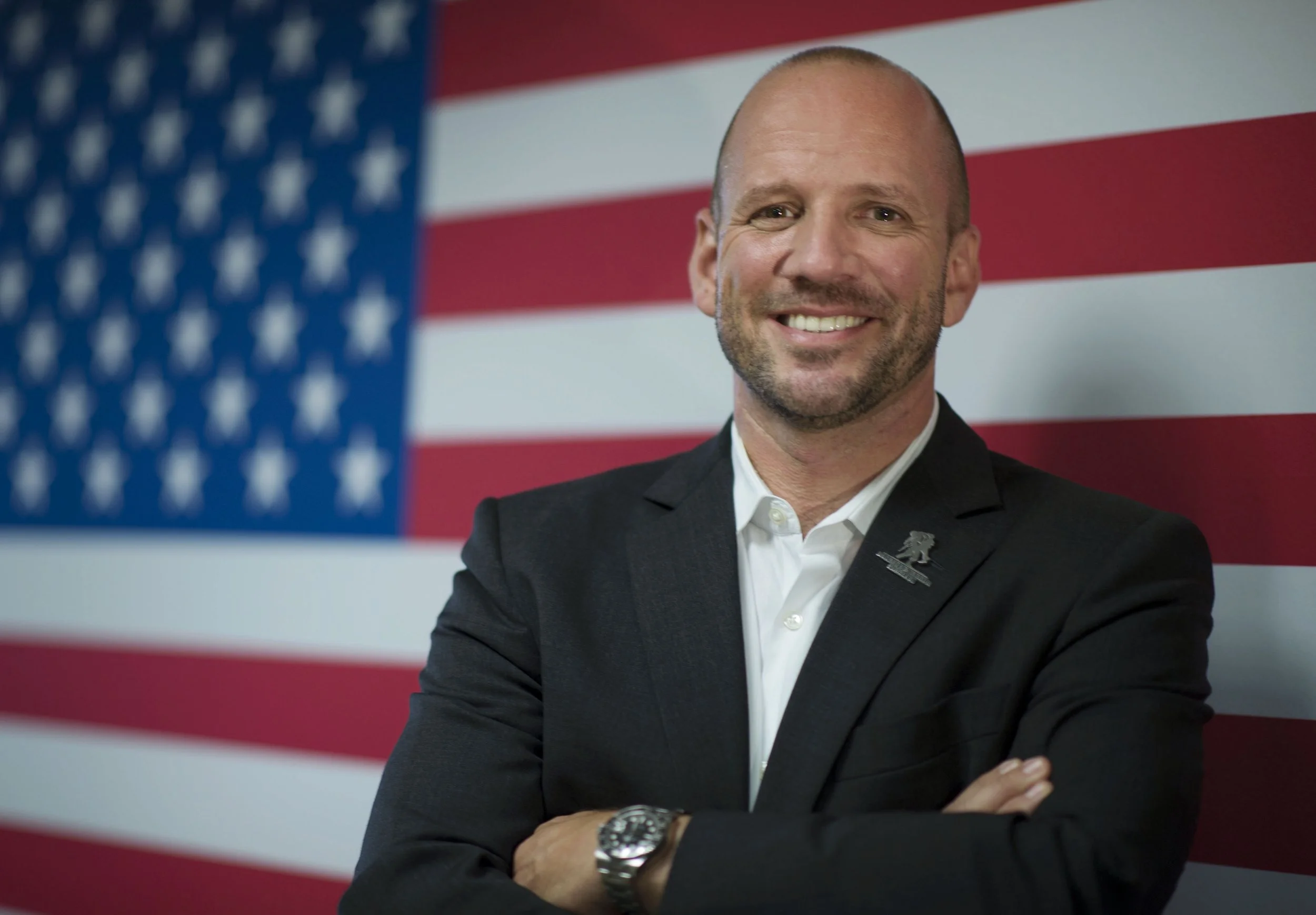 Dan Nevins is in a black suit and white shirt, standing in front of the American flag.
