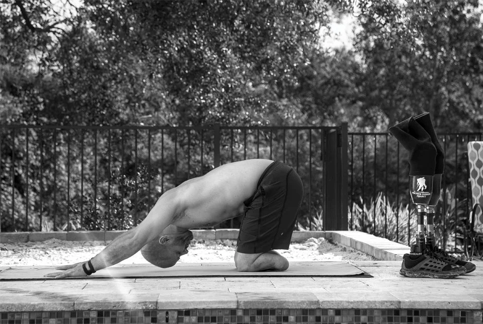 Dan Nevins practicing yoga outdoors, kneeling with his head down and arms extended, on a mat near a fence and trees.