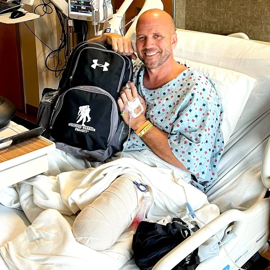 Dan Nevins in a hospital bed smiling, holding a backpack with an Under Armour logo and a Wounded Warrior Project logo.